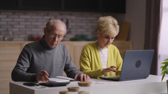退休的男人和女人一起坐在家里的厨房里计算费用，老人用计算器计数，老太太用笔记本电脑
