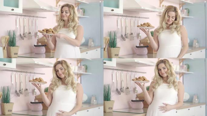 金发碧眼的孕妇拿着盘子，拿着健康的甜饼干，对着镜头微笑。年轻美丽的高加索人的肖像站在家里的厨房里。生