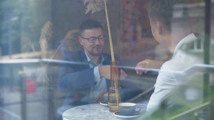 两个高加索商人坐在咖啡馆里聊天。自信的年轻男性企业家在食堂讨论生意。在玻璃后面射击。创业理念。