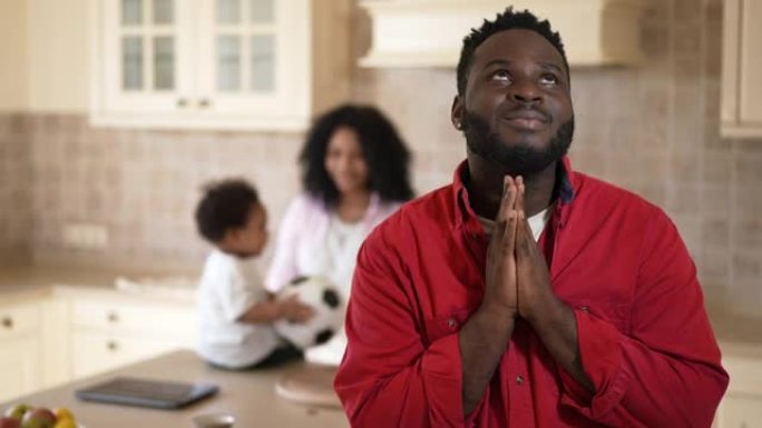 快乐兴奋的非洲裔美国年轻人祈祷感谢上帝给家人，模糊的妻子和儿子在后台玩耍。英俊的父亲丈夫在室内厨房的
