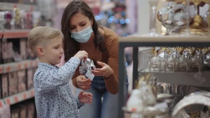 一位戴着医用口罩的母亲和她的儿子在购物中心选择圣诞节的家居装饰品。观看圣诞树玩具。冠状病毒流行病