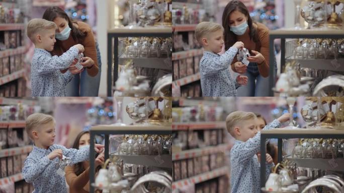一位戴着医用口罩的母亲和她的儿子在购物中心选择圣诞节的家居装饰品。观看圣诞树玩具。冠状病毒流行病