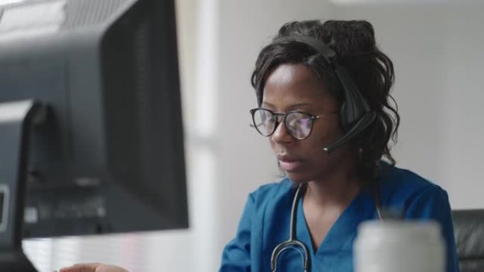 非洲女医疗助理穿着白大褂，耳机视频在电脑上给远方病人打电话。医生使用虚拟聊天计算机应用程序与客户交谈
