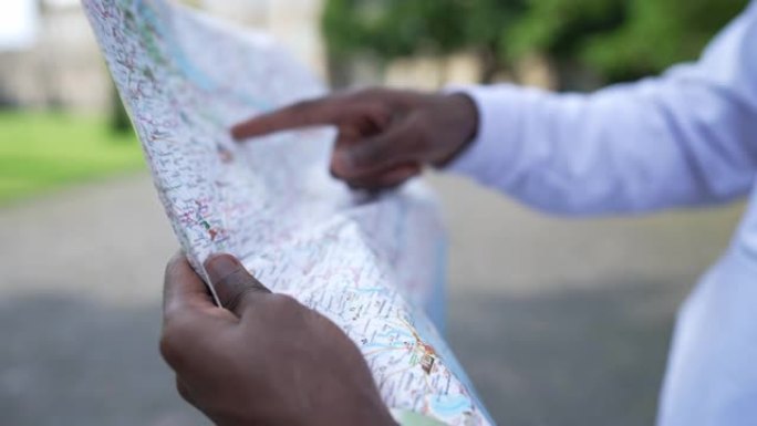 男性非裔美国人的手指向纸质地图搜索方向。无法识别的年轻人选择春季夏日站在户外的路线。出国旅游的游客。