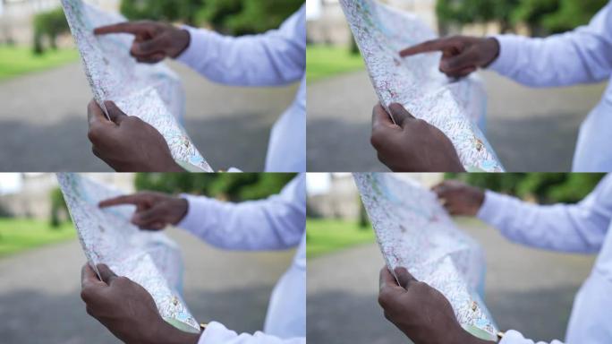 男性非裔美国人的手指向纸质地图搜索方向。无法识别的年轻人选择春季夏日站在户外的路线。出国旅游的游客。