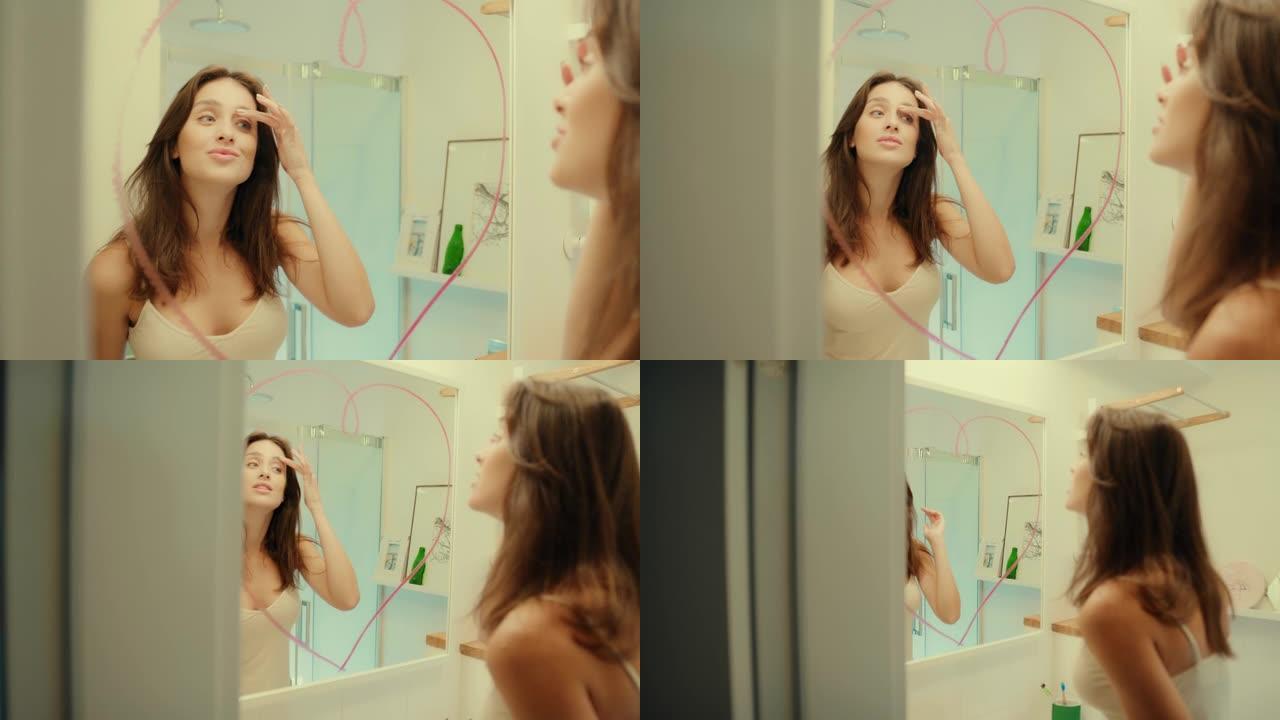 诱人的黑发女人享受她的倒影。在镜子上画口红心。早上浴室例行公事