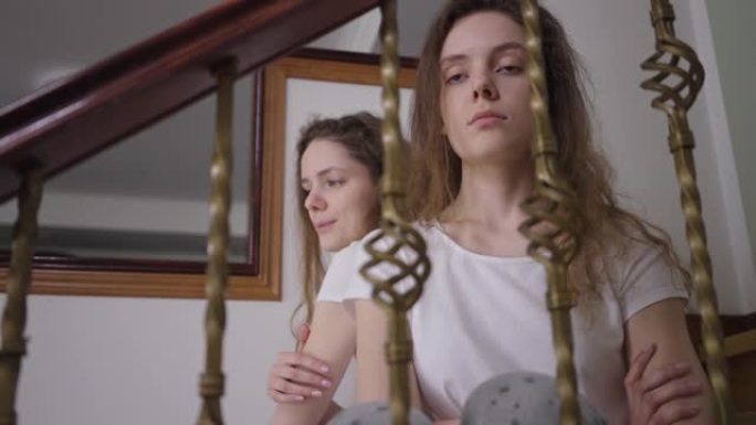沮丧的白人年轻女子坐在楼梯上，双胞胎妹妹在背景上说话。悲伤美丽的千禧一代兄弟姐妹与姐姐争吵，思念叹息