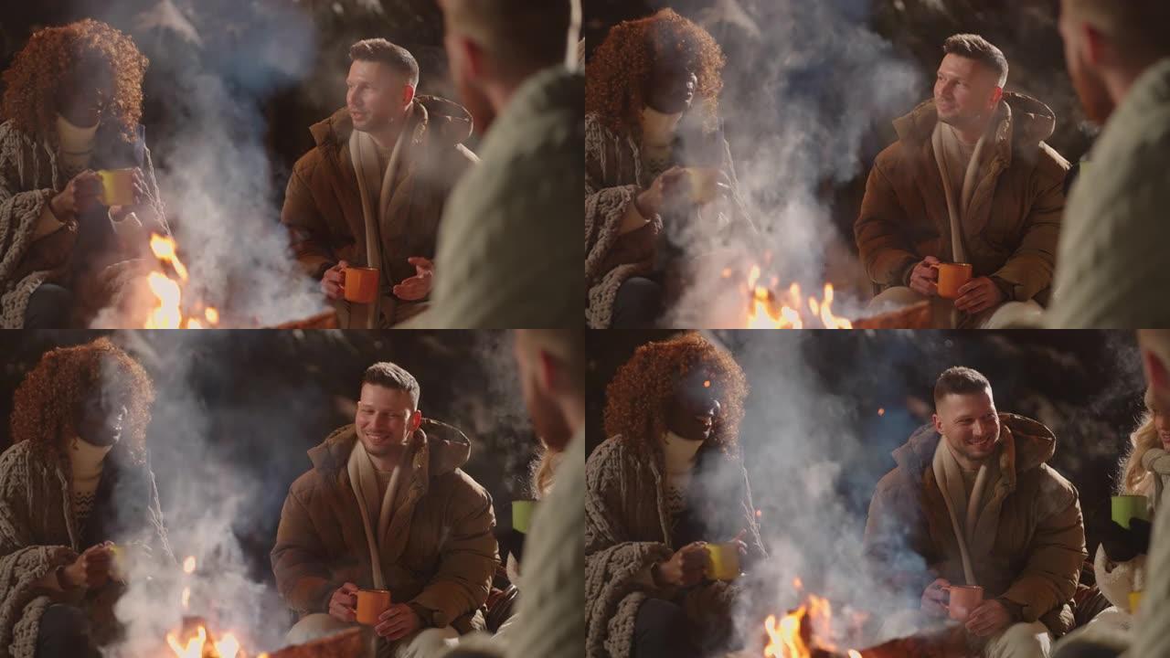 黑人和高加索人的朋友在冬天的夜晚在篝火旁变暖，喝热酒和聊天
