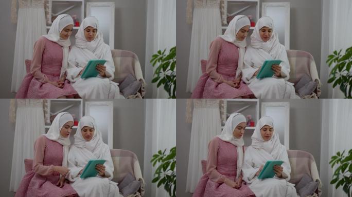婚礼当天早上，迷人自信的中东女性戴着白色头巾在室内平板电脑上浏览社交媒体。两个积极的穆斯林妇女坐在沙