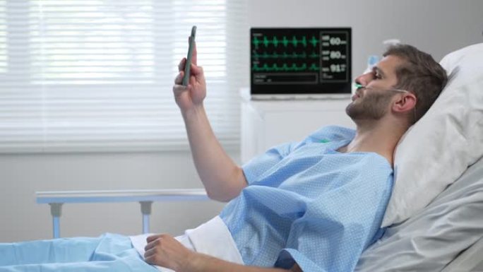 一名年轻的男性诊所患者躺在医院的病床上，并通过手机通过视频链接与家人和朋友通话。