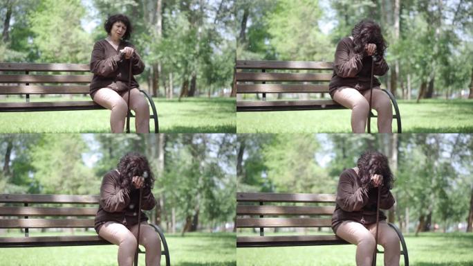 在春季或夏季公园，沮丧的高加索高级妇女在拐杖上哭泣。沮丧的孤独女性退休人员坐在长凳上，花粉飞来飞去。