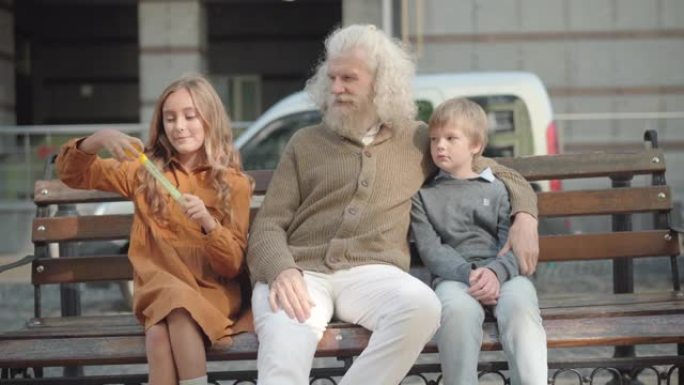 放松的高加索祖父和孙子坐在长椅上，孙女吹肥皂泡。快乐放松的多代家庭在户外享受周末的肖像。