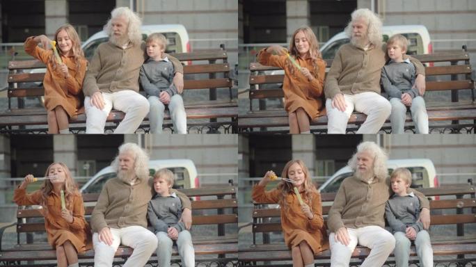 放松的高加索祖父和孙子坐在长椅上，孙女吹肥皂泡。快乐放松的多代家庭在户外享受周末的肖像。