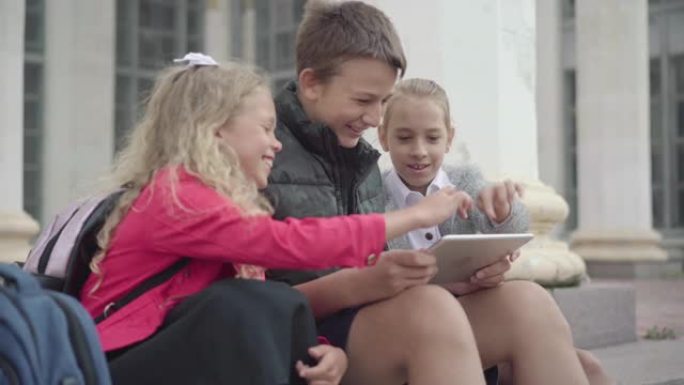 三个开朗的孩子同学在平板电脑上使用社交媒体并聊天。快乐的高加索男生和女学生在休息时笑着聊天的侧视图。