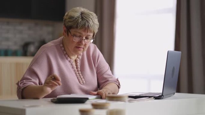 戴眼镜的退休妇女正在用计算器计算水电费，独自一人坐在家庭厨房里，家庭主妇正在控制财务状况