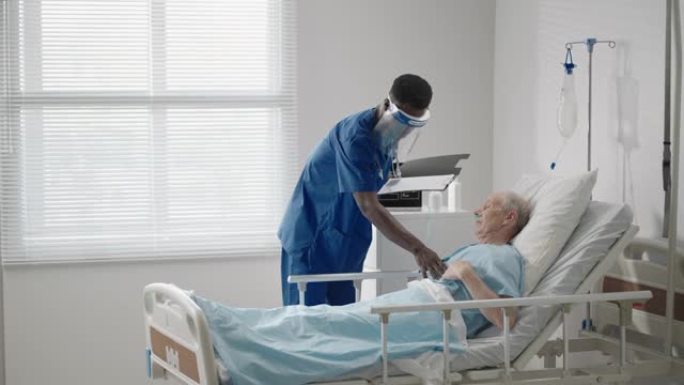 一名黑人男性外科医生正在和一名躺在医院病床上的老年男性患者交谈。一名女性心脏病专家传染病专家正在诊所