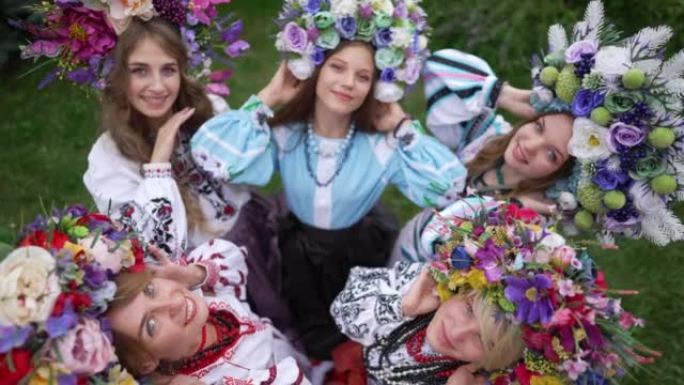 顶视图迷人的乌克兰年轻女性戴着花圈和民族服装抬头看着相机微笑。在夏季春季公园摆姿势的美丽女士的高角度
