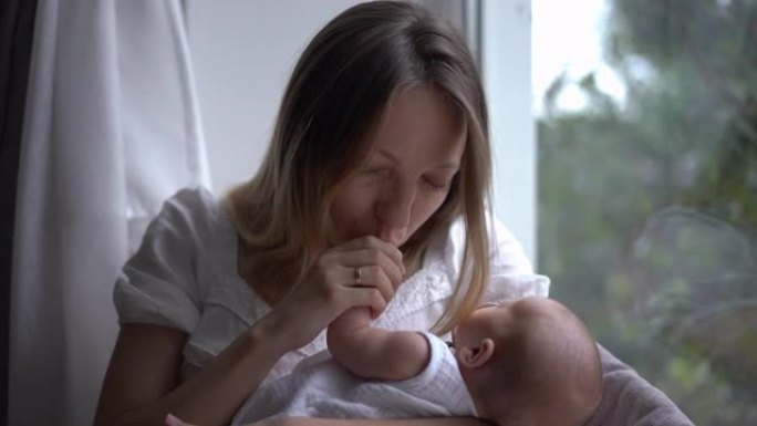 白人妇女在室内微笑着亲吻坐在窗台上的新生婴儿的小手。爱快乐的母亲欣赏可爱的小婴儿儿子的肖像，享受休闲
