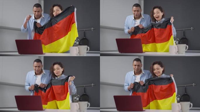 快乐的跨种族夫妇与德国国旗为足球队欢呼，在厨房的笔记本电脑上观看在线比赛。年轻的非洲裔美国男子和白人