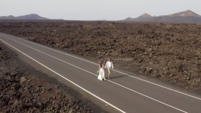 空中无人机拍摄的新婚新娘和新郎在火山岛上的一条空旷的道路上行走，背后是群山