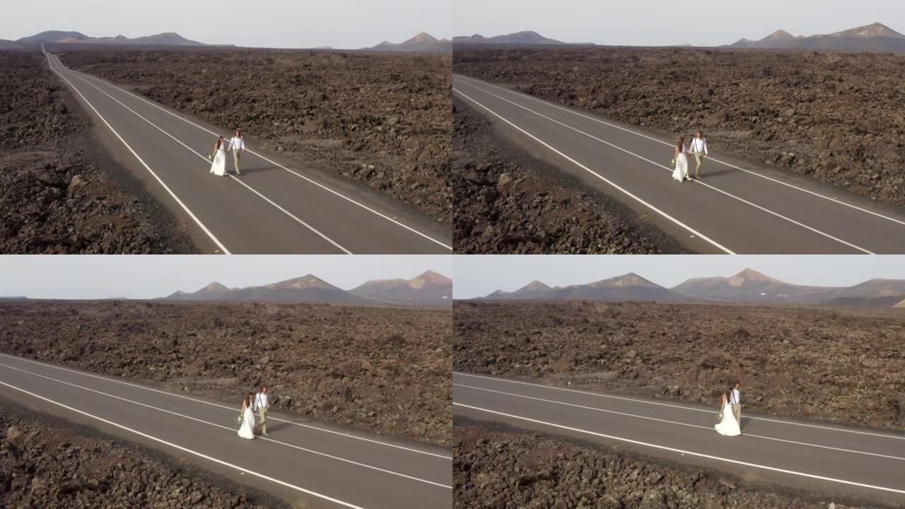 空中无人机拍摄的新婚新娘和新郎在火山岛上的一条空旷的道路上行走，背后是群山