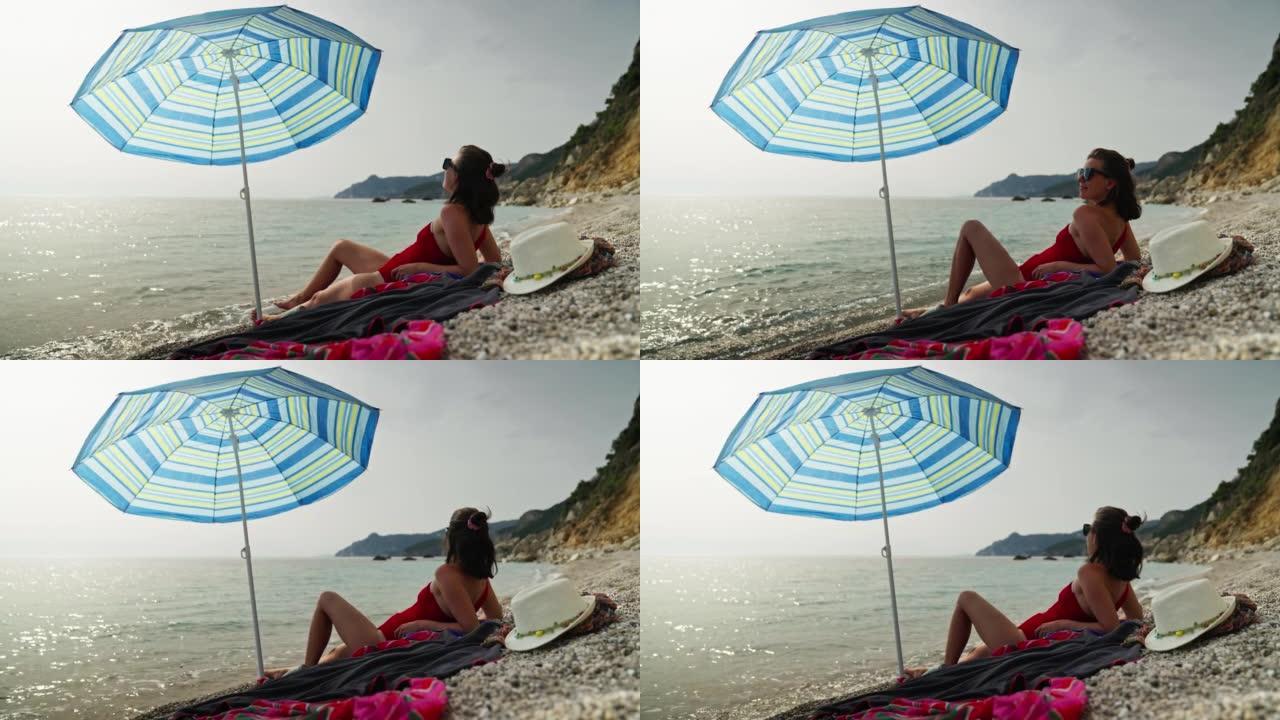 戴着墨镜的女人在石滩上在沙滩伞下放松。享受大海
