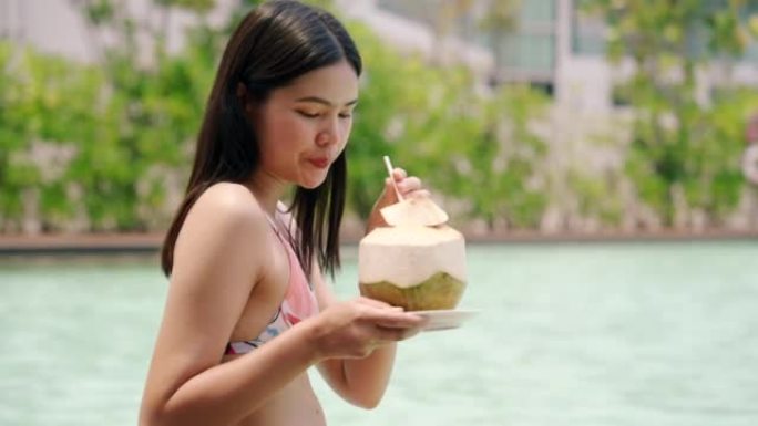 穿着比基尼泳装的亚洲女孩坐在泳池边喝椰子水放松玩手机。