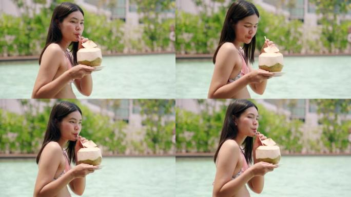 穿着比基尼泳装的亚洲女孩坐在泳池边喝椰子水放松玩手机。