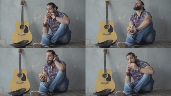 宽镜头正面的年轻沮丧的男子坐在地下交叉吉他和吃。白人音乐家乞讨后吃午饭的肖像。