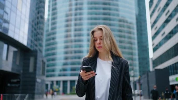 在摩天大楼的背景下，一个年轻的女人走在大都市的街道上，手里拿着手机，一边看新闻一边做生意