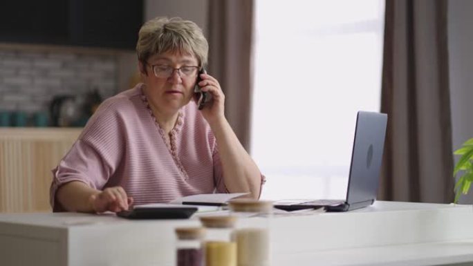 老太太正在打电话，在家工作，独自坐在厨房里，使用笔记本电脑和计算器