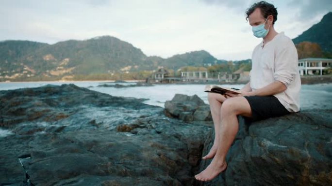 一个戴着面具的男人坐在海洋附近的岩石上看书的特写镜头