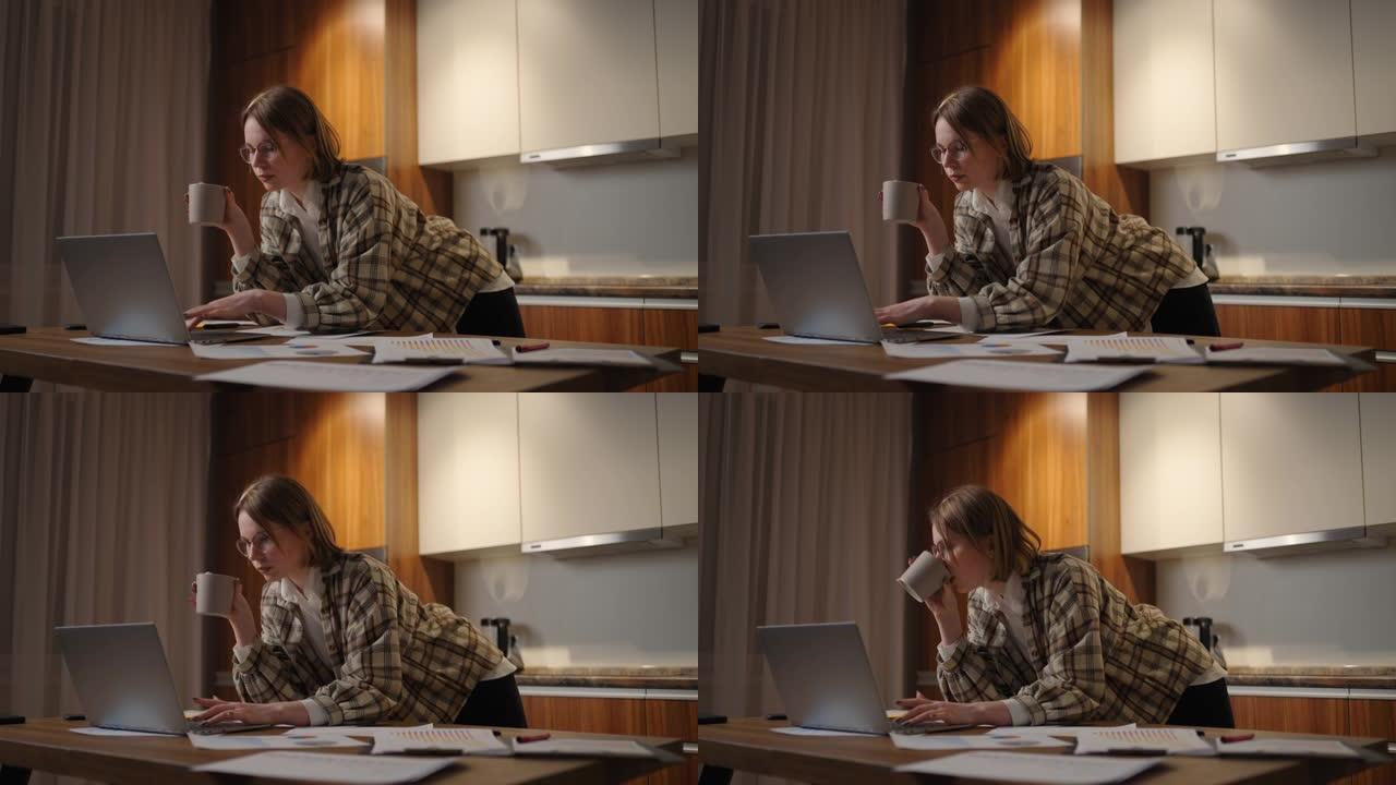 一位年轻漂亮的女商人在家工作，喝杯咖啡，看着站在桌子旁的笔记本电脑屏幕。查看邮件和喝咖啡。在家远程工