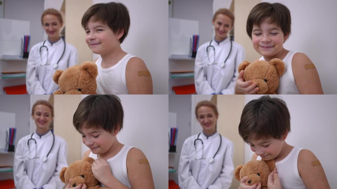 快乐的高加索男孩在冠状病毒疫苗接种中心玩泰迪熊，看着相机微笑。快乐可爱的孩子的肖像，肩膀上有医疗贴片