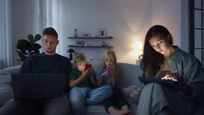 黑暗中的家庭迟来的粉丝坐在潜水上，妈妈在平板电脑上画画，爸爸在笔记本电脑上工作，孩子们看着手机的屏幕