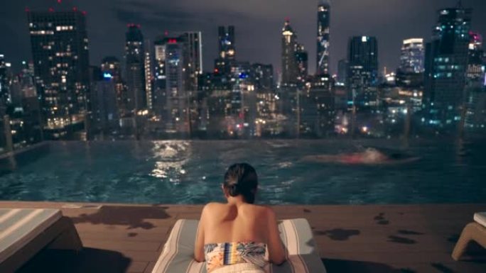 晚上，一名亚洲男子游泳和女子拿着手机坐在一栋高层建筑的游泳池里，俯瞰着这座城市。