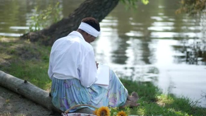 年轻专注的非洲裔美国妇女坐在河岸上，以慢动作写日记。吸收无忧无虑的女士计划未来在户外享受周末休闲的宽