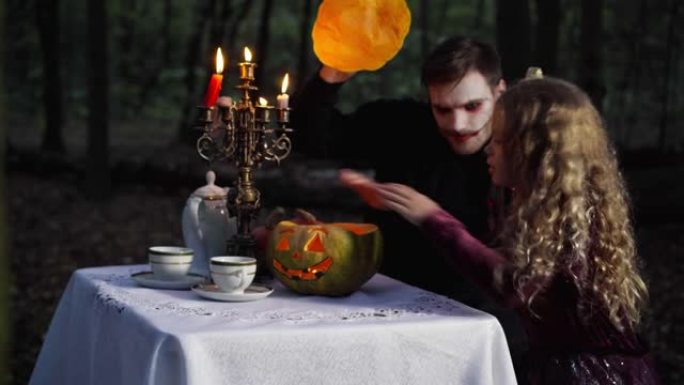 万圣节期间，小漂亮女孩和穿着吸血鬼服装的男人坐在黑暗的森林里，用蜡烛打开蔬菜。快乐的高加索兄妹庆祝。