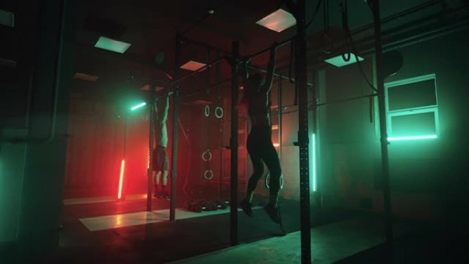 健身房里的两个男人和一个女人在黑暗的霓虹灯下一起在单杠上做引体向上。健身和训练的氛围