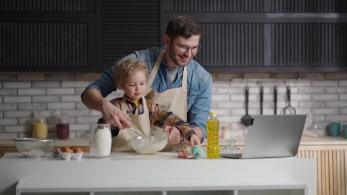 年轻的父亲和小儿子正在通过视频教程做饭，在笔记本电脑上观看视频并鞭打煎饼的面团