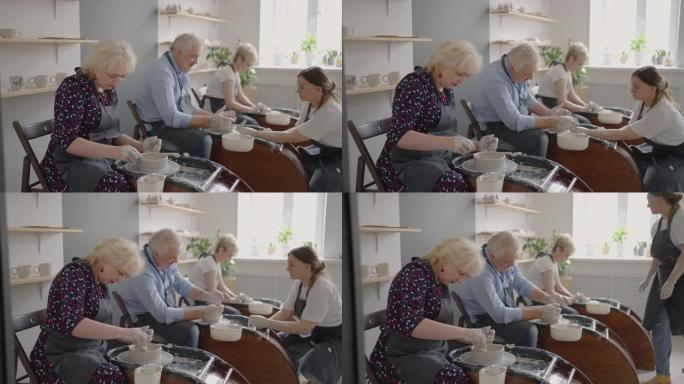 中年陶瓷艺术家教学小组老年白人妇女和高级男子如何在艺术工作室的办公桌上楔入粘土。人们喜欢在工作中交谈