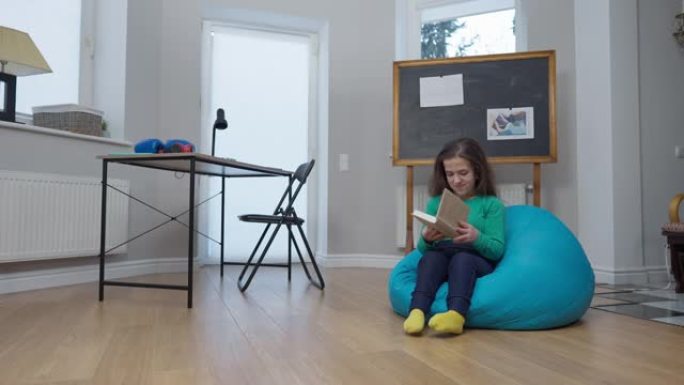 宽大的客厅，配有书桌和黑板，高加索小人坐在舒适的书包椅子上打开书。积极微笑的美女在家室内阅读。