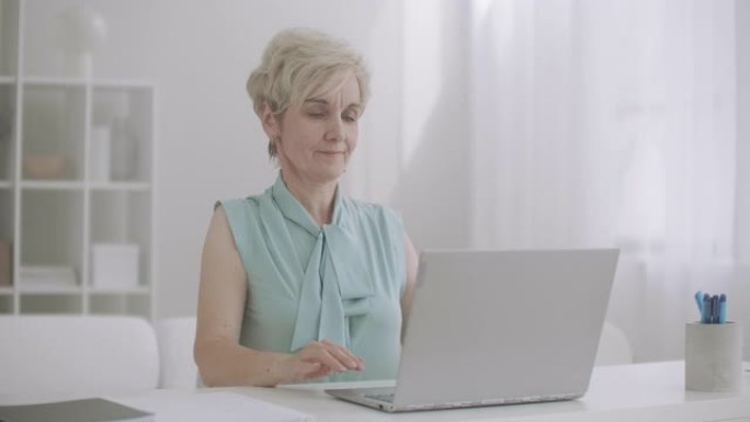 患有骨软骨病的退休妇女在使用笔记本电脑，坐在桌子旁并在键盘上打字时伸展手和脖子