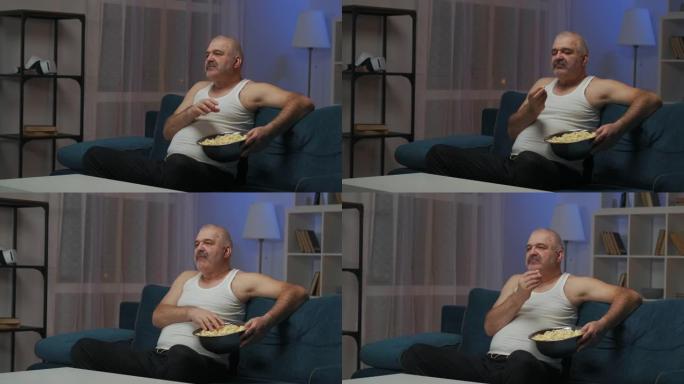 一个男人晚上看电视。他坐在沙发上。吃大铁盘子里的爆米花。摄像机从右向左移动