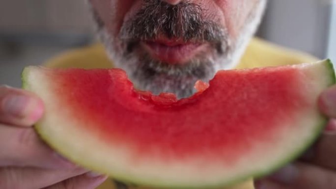 特写红西瓜片，面目全非的大胡子男人在慢动作中吃美味多汁的浆果。白人男子在夏天在室内享受甜食的味道。