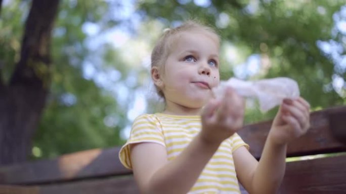 小女孩吃完后用餐巾纸擦脸，桌上放着奶昔。可爱的小女孩坐在公园长椅上，用纸巾擦脸的特写镜头。