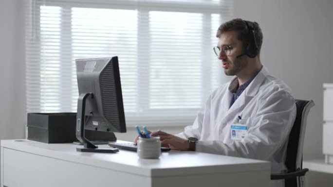 在医院工作的带耳机和计算机的男医生或护士。医疗操作员与耳机咨询患者的特写。在药品呼叫中心工作的医务人