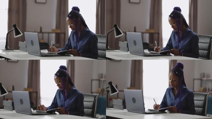 女老板在办公室工作，黑人妇女在笔记本上做笔记，坐在笔记本电脑旁，在笔记本电脑上写待办事项清单
