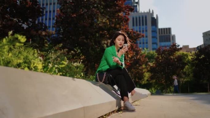 休闲风格的年轻韩国女性在城市公园休息，并通过现代智能手机上网