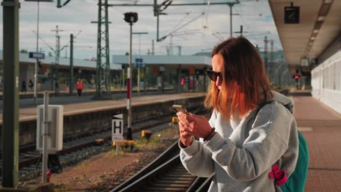 女旅行者站在空荡荡的火车站平台上，看着智能手机显示屏，搜索火车的到达时间。旅行的概念。站在车站的背包